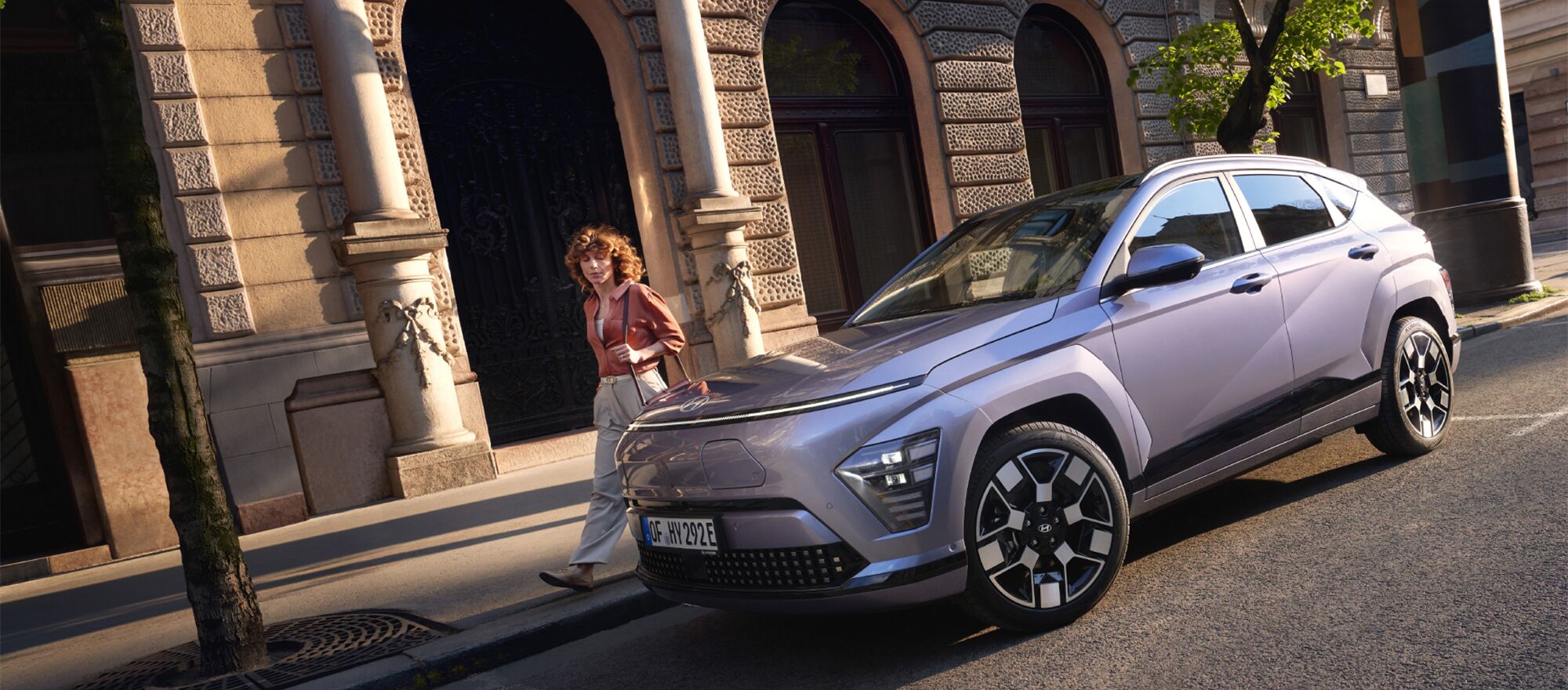 Ein parkender Hyundai KONA Elektro vor einem Gebäude und ein Frau die hinter dem Auto läuft