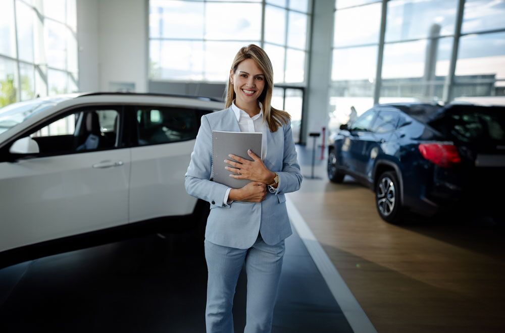 Eine Frau steht mit einer Mappe in der Hand lächelnd im Verkaufsraum eines Autohauses