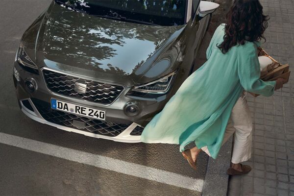 Der Kühlergrill des SEAT Arona an dem eine Frau mit ihren Einkäufen vorbei läuft
